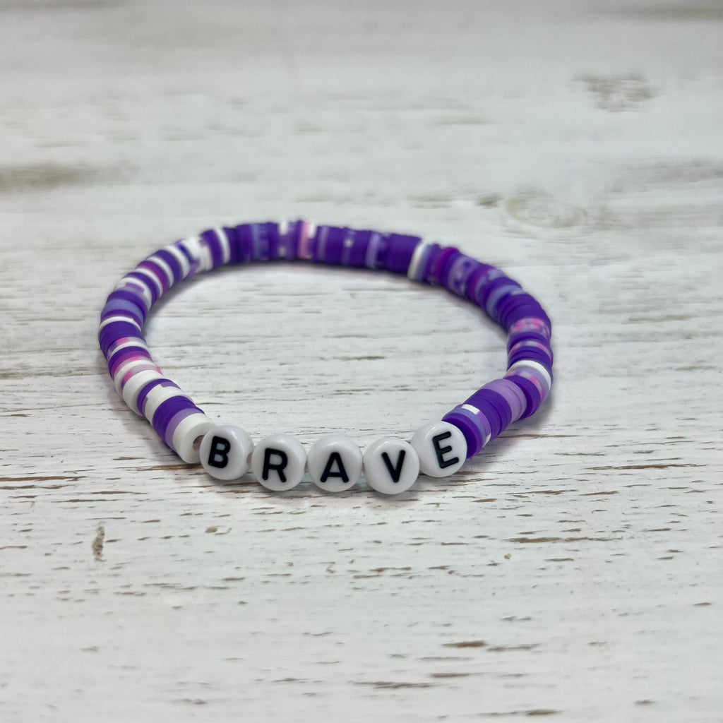 Bracelet with purple and white beads spelling 'BRAVE' on a wooden surface