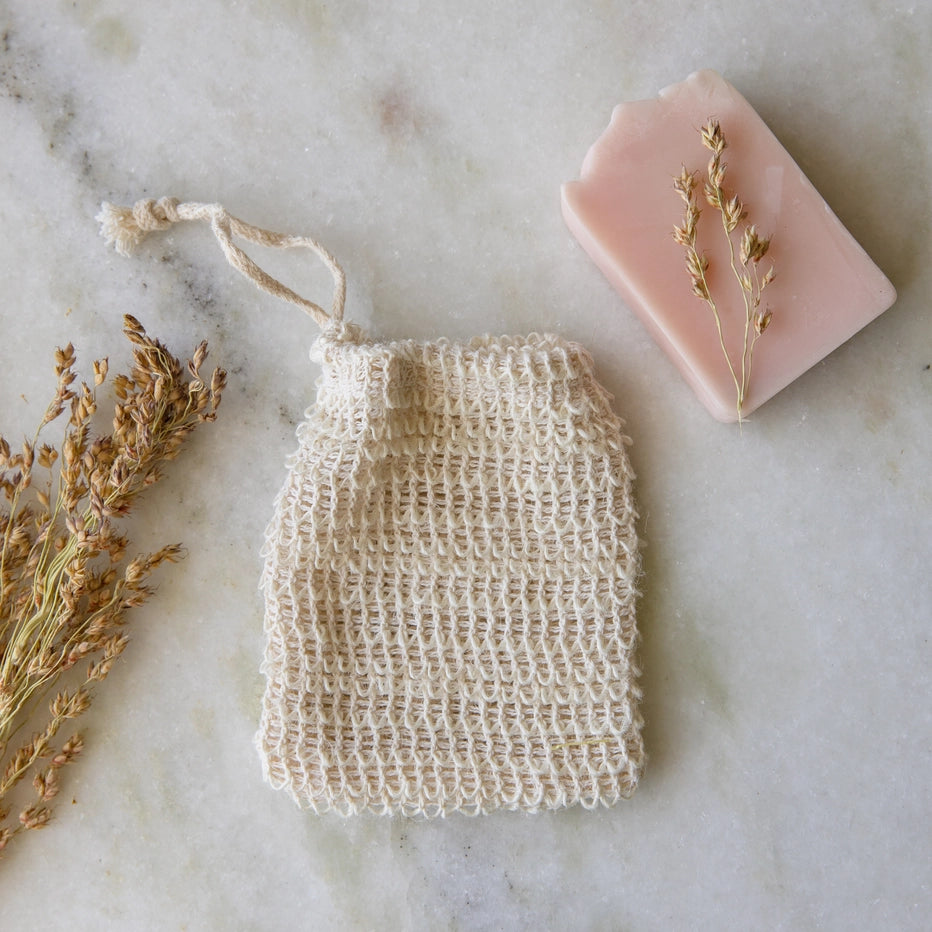 Natural soap bag, pink bar of soap, and dried herbs on a marble surface