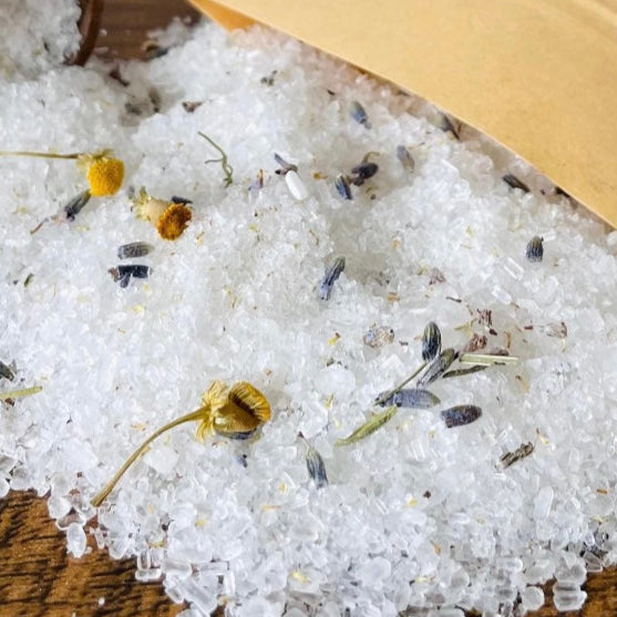 Bath salt with herbs on a wooden surface next to an open bag labeled 'Bath Salt'.