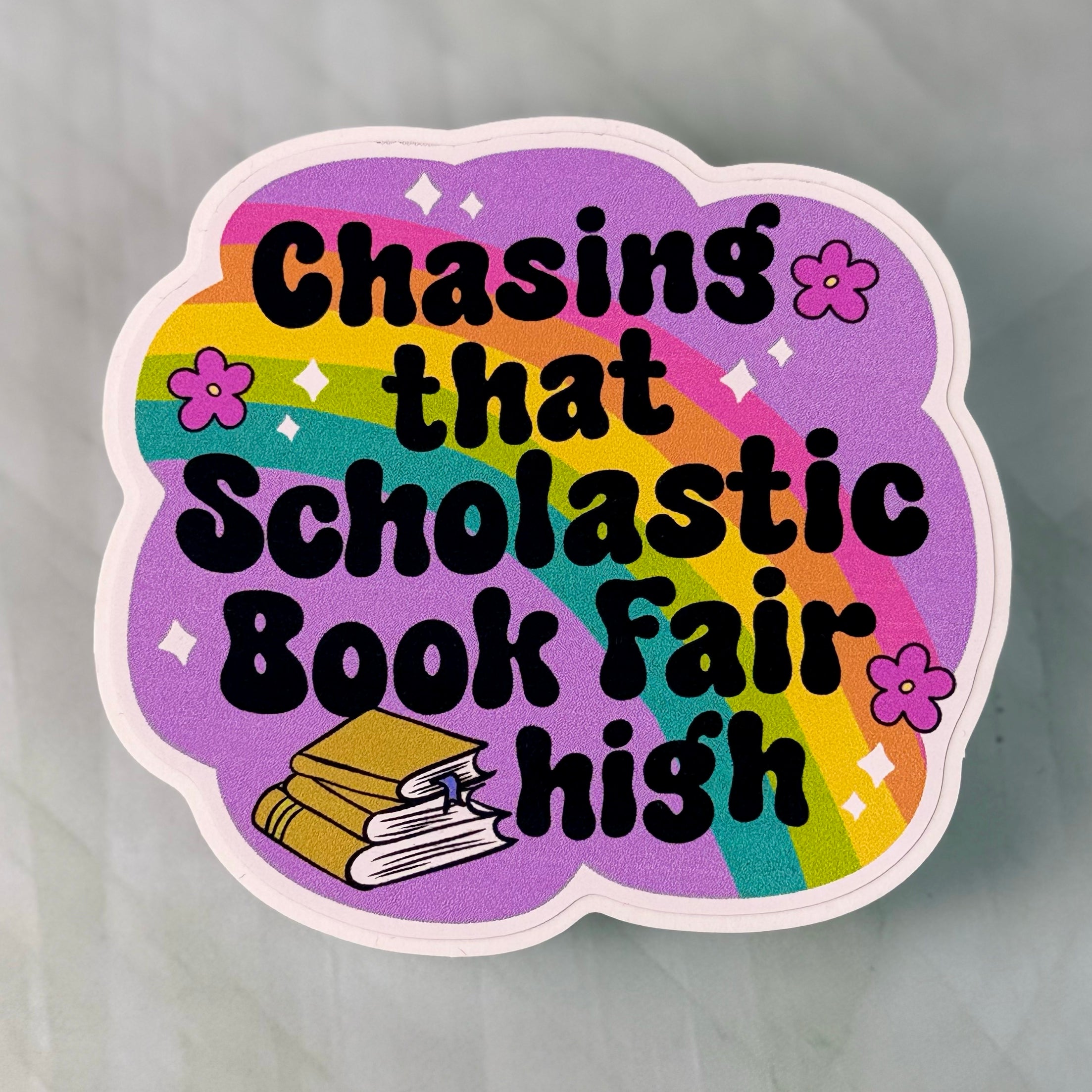 Sticker with text 'Chasing that Scholastic Book Fair high' on a purple background with rainbow and flowers.