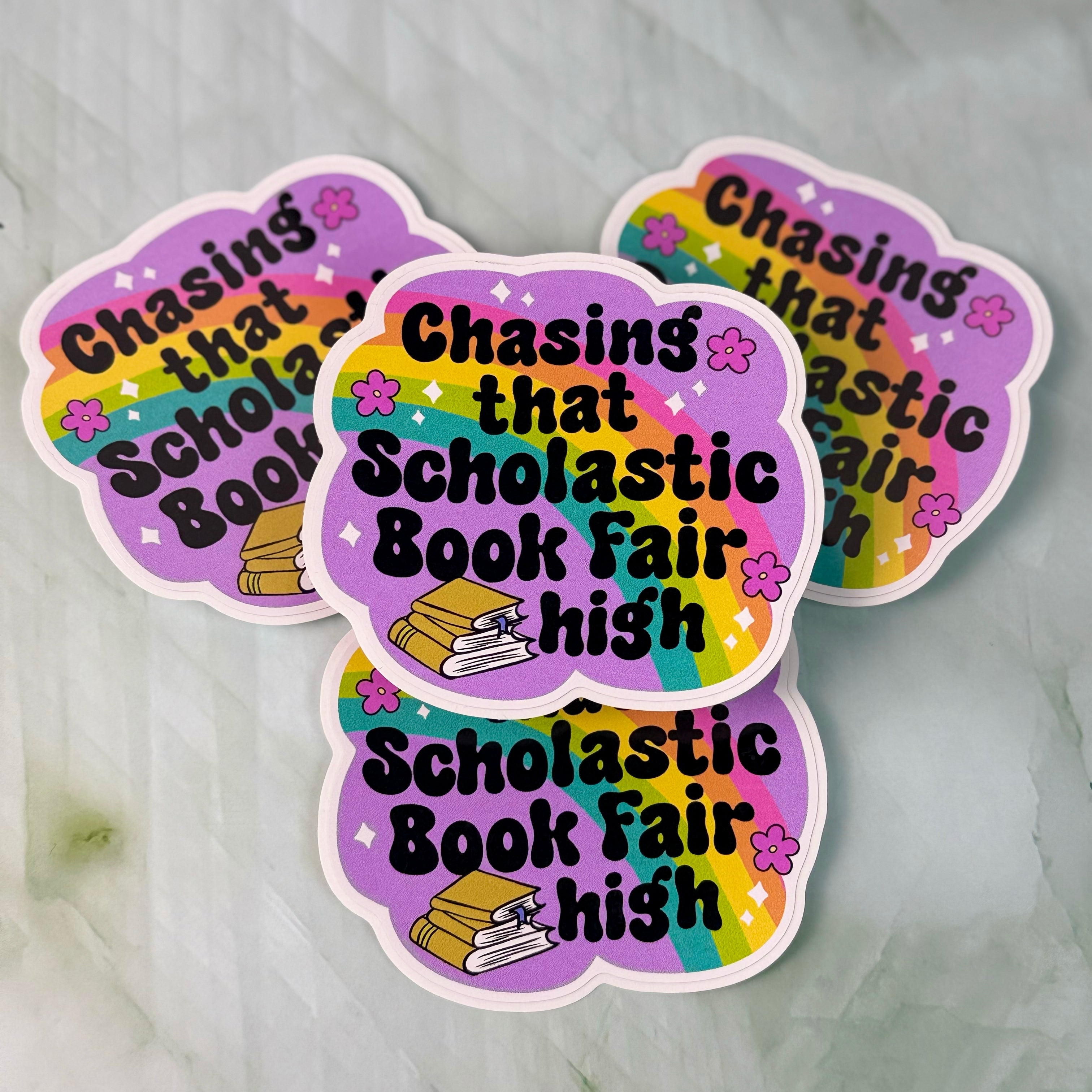 Three colorful stickers with text 'Chasing that Scholastic Book Fair high' on a marble surface.