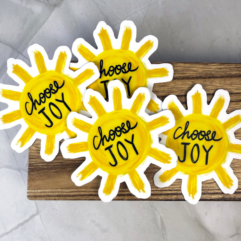 Four yellow sun-shaped stickers with 'Choose Joy' text on a wooden block.