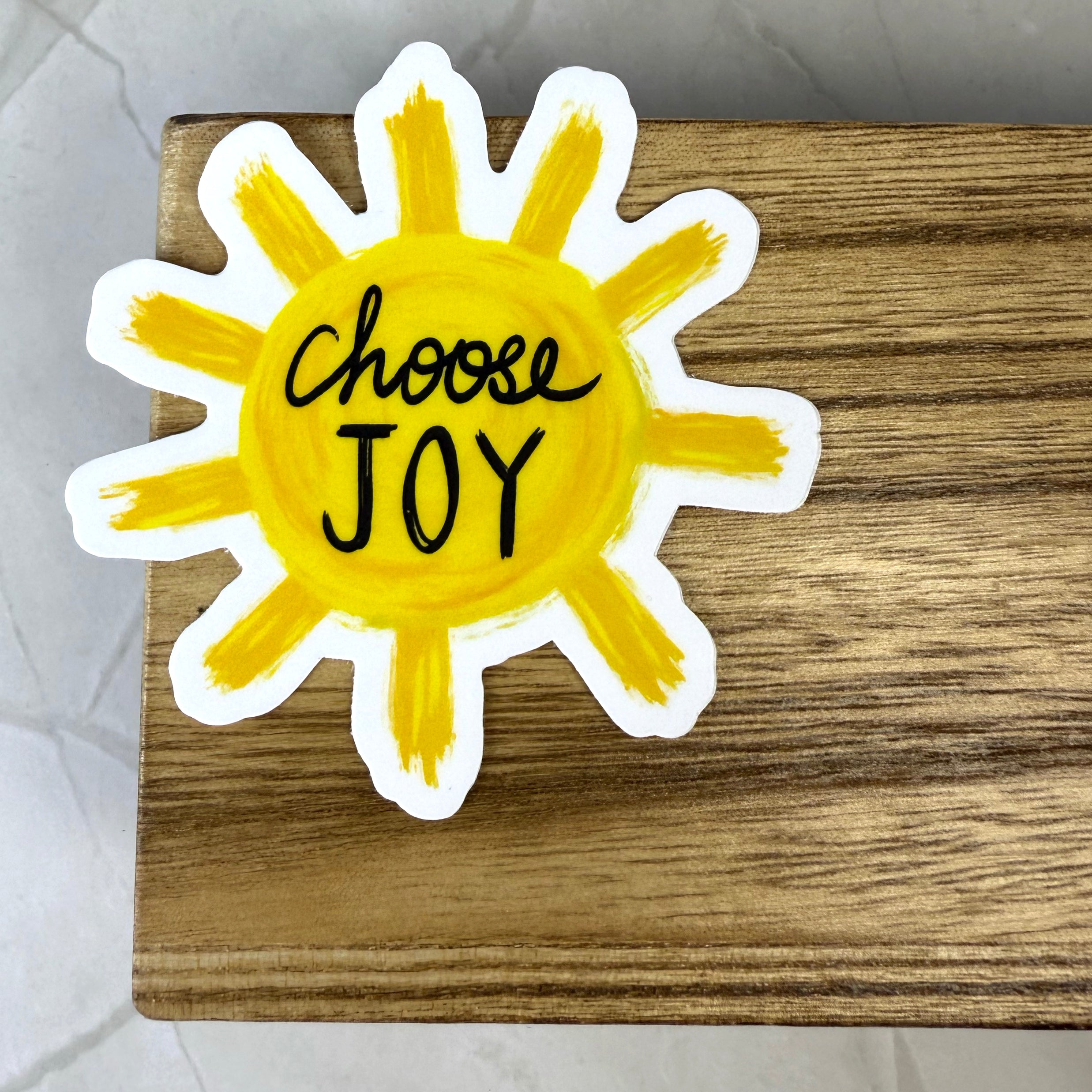 Yellow sun-shaped sticker with 'Choose Joy' text on a wooden surface