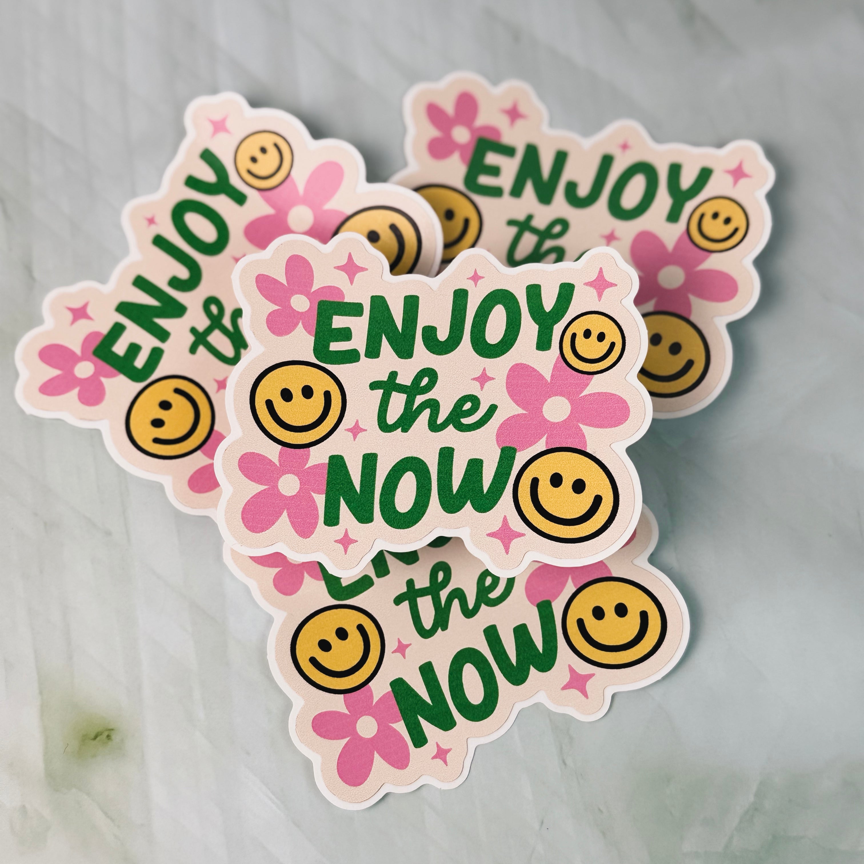Four stickers with 'Enjoy the Now' text and smiley faces on a light gray background