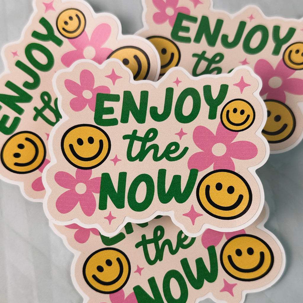 Sticker with 'Enjoy the Now' text and smiley faces on a pink background