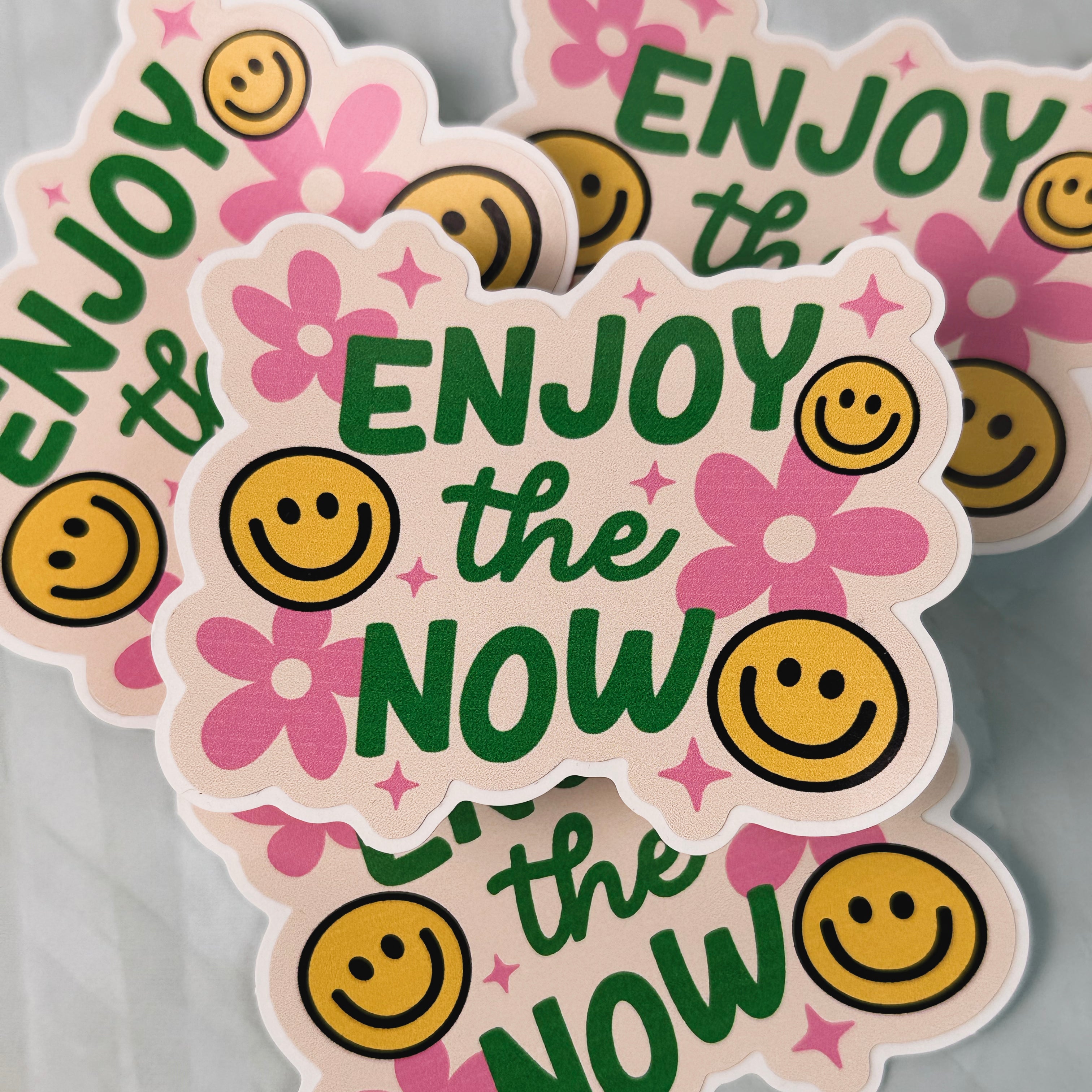 Sticker with 'Enjoy the Now' text and smiley faces on a pink background