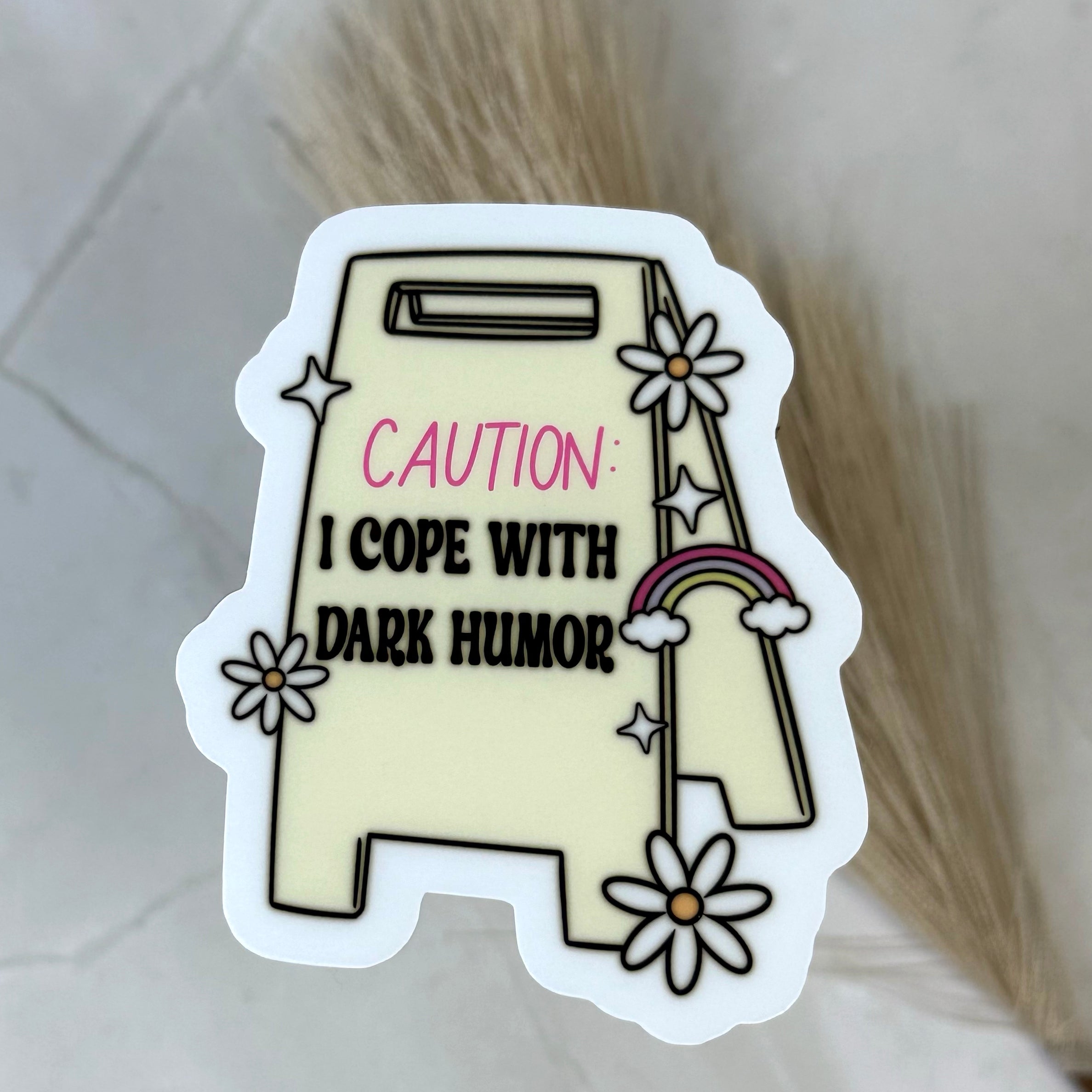 Sticker with text 'CAUTION: I COPE WITH DARK HUMOR' on a light background