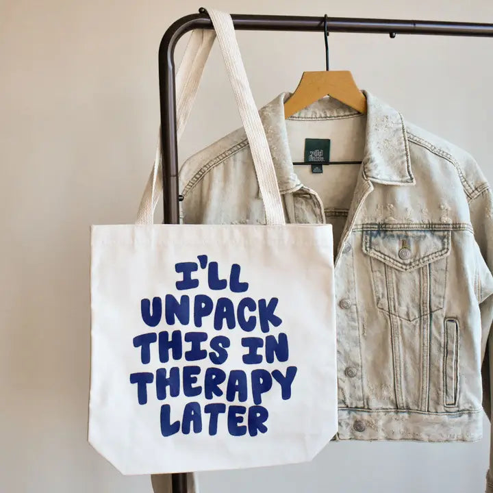 Tote bag with 'I'll unpack this in therapy later' text hanging on a rack with a denim jacket.