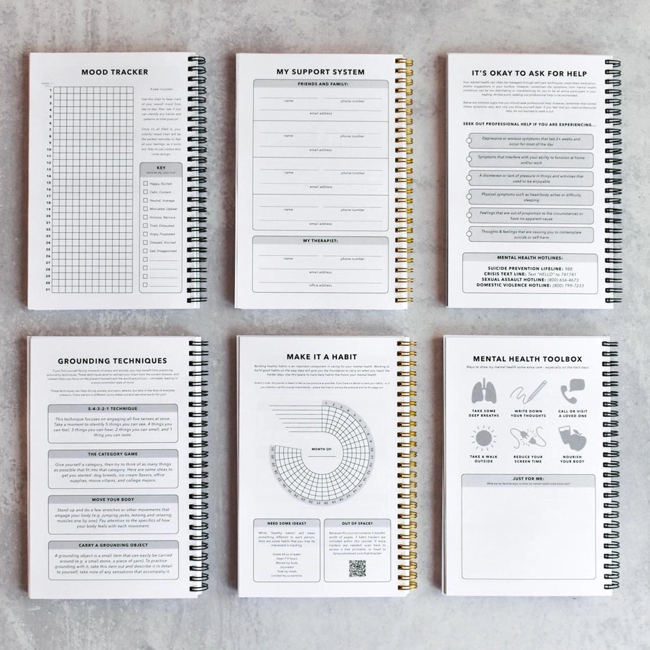 Six spiral-bound journals with mental health tools and trackers on a gray surface