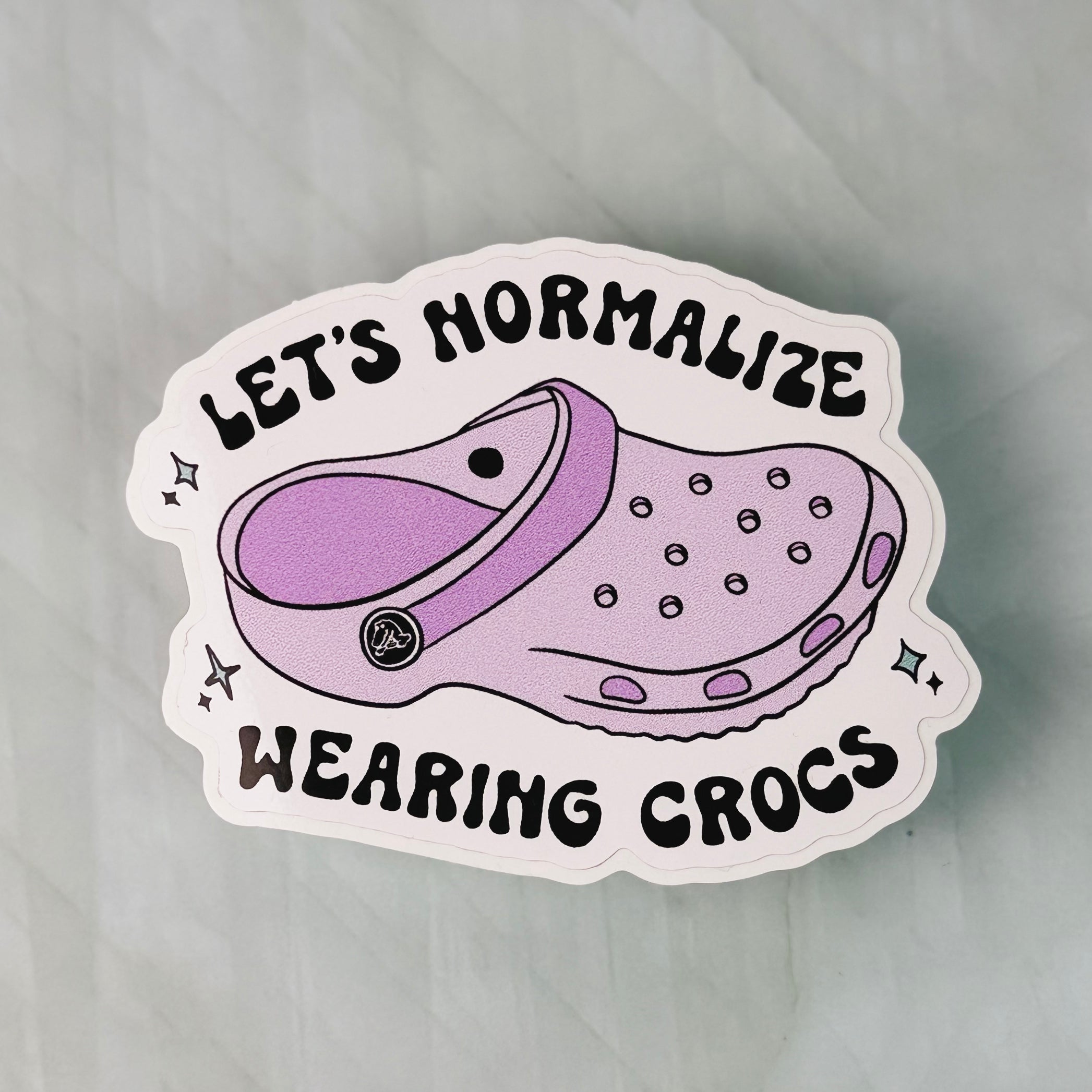 Sticker with a purple croc shoe and text 'Let's Normalize Wearing Crocs' on a light gray background
