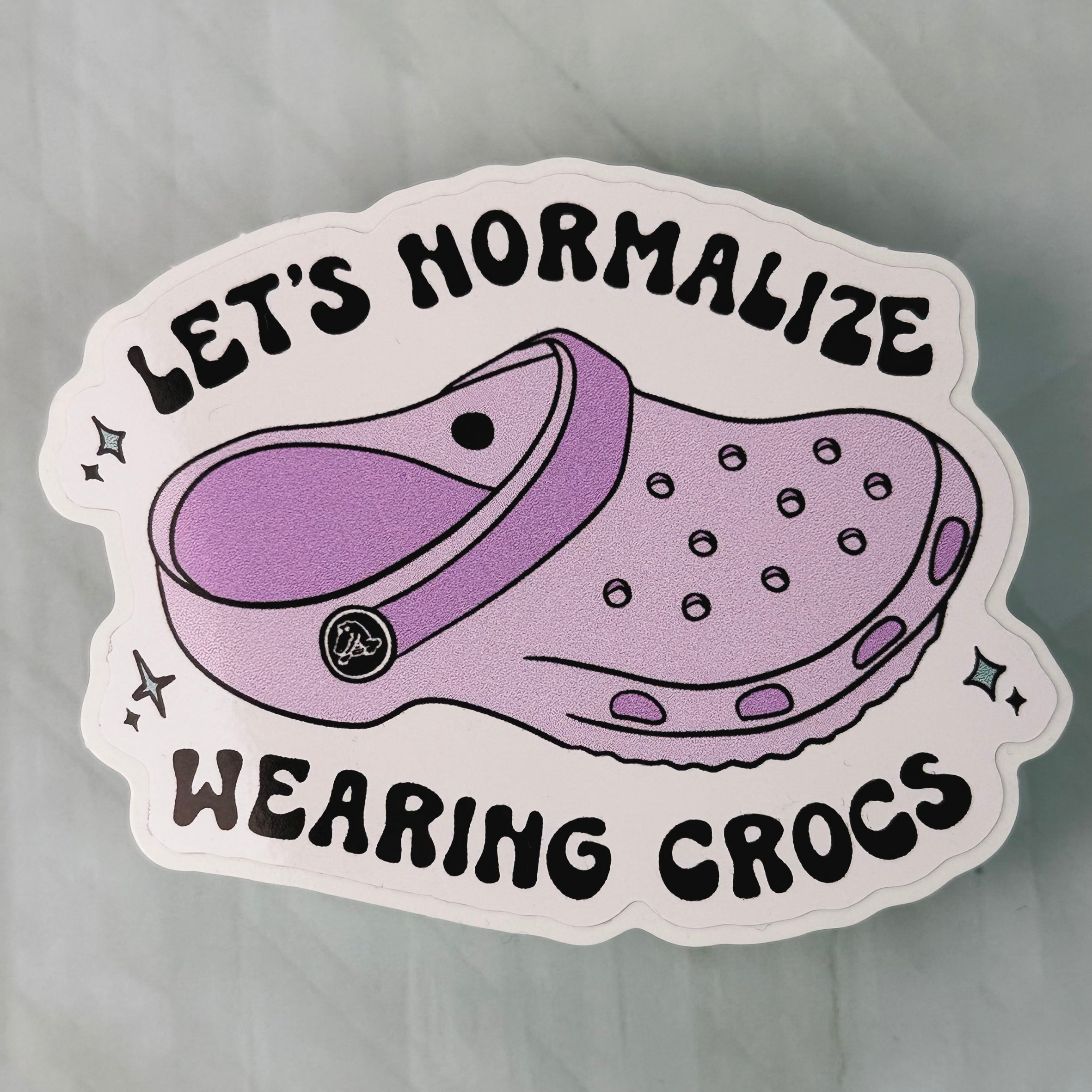 Sticker with a purple croc shoe and text 'Let's Normalize Wearing Crocs' on a gray background