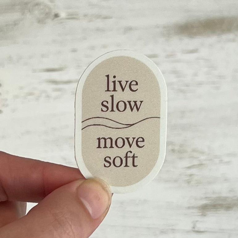 Sticker with 'live slow move soft' text held by a hand on a wooden surface
