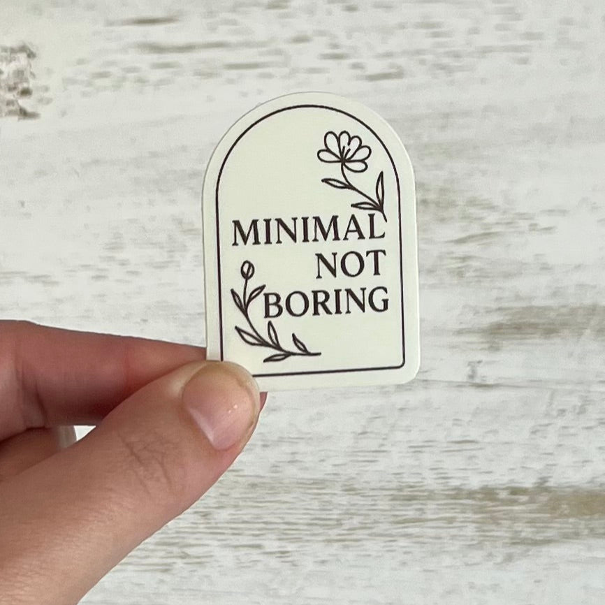 Sticker with 'Minimal Not Boring' text held by a hand on a wooden surface