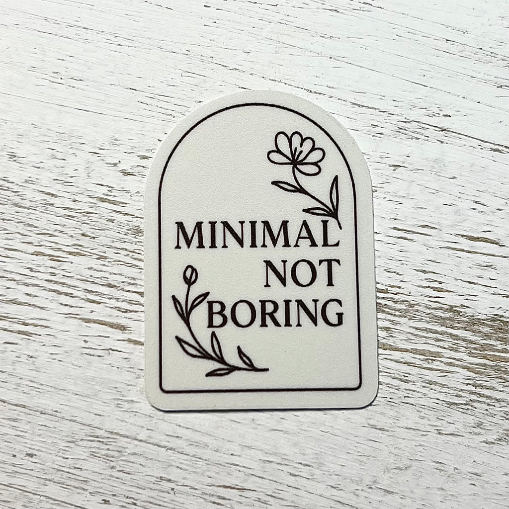 Sticker with 'Minimal Not Boring' text on a white wooden surface