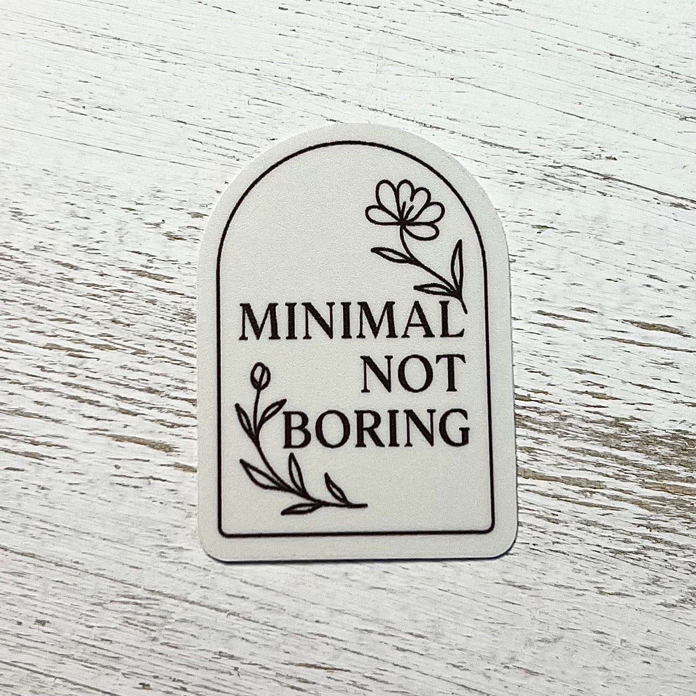 Sticker with 'Minimal Not Boring' text on a white wooden surface