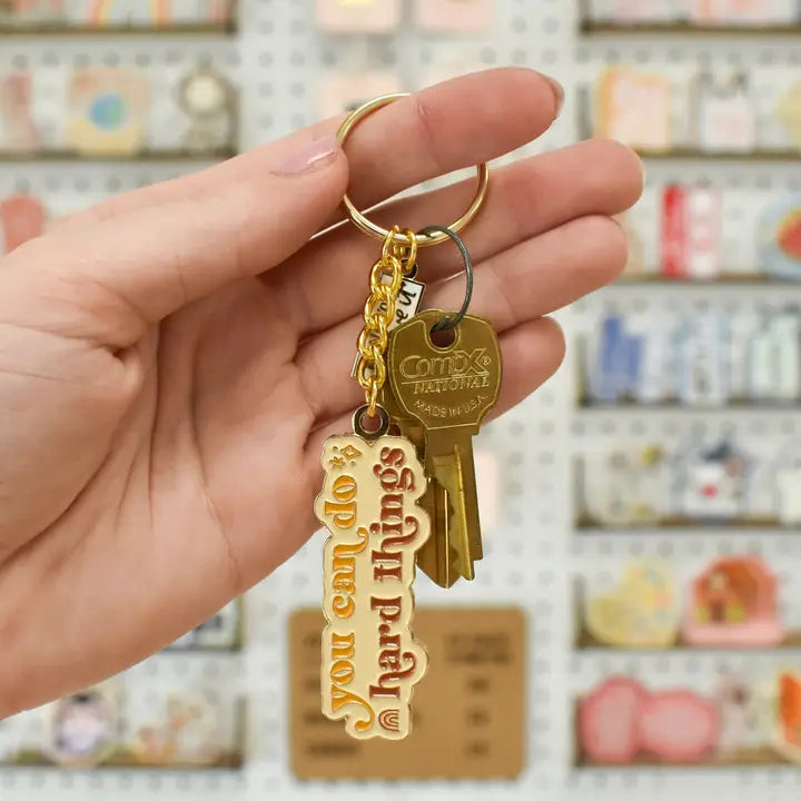 Hand holding a key with a decorative tag saying 'You can do hard things' in front of a store shelf.